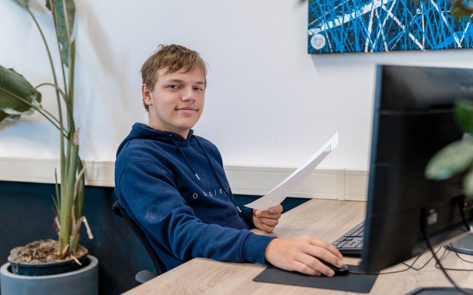 Jongen zit met papier in zijn linkerhand te werken achter computer.