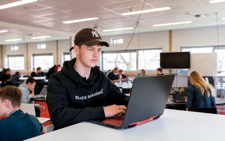 Student Software Developer zit aan hoge tafel achter laptop in een klaslokaal vol met medestudenten