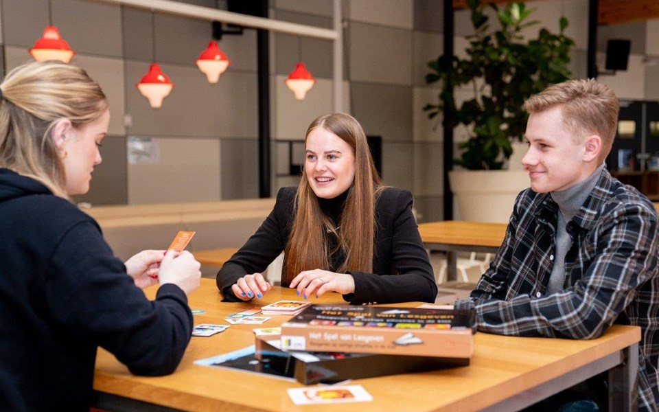 Student onderwijsassistent leest kaartje voor aan medestudenten tijdens spel aan tafel. 