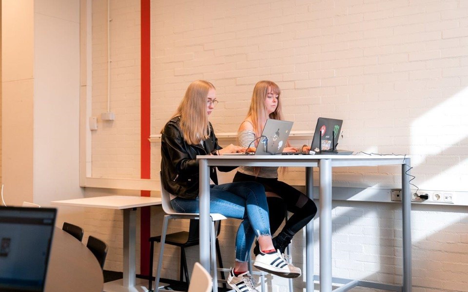 Twee studenten ICT zitten naast elkaar aan hoge tafel achter hun laptop