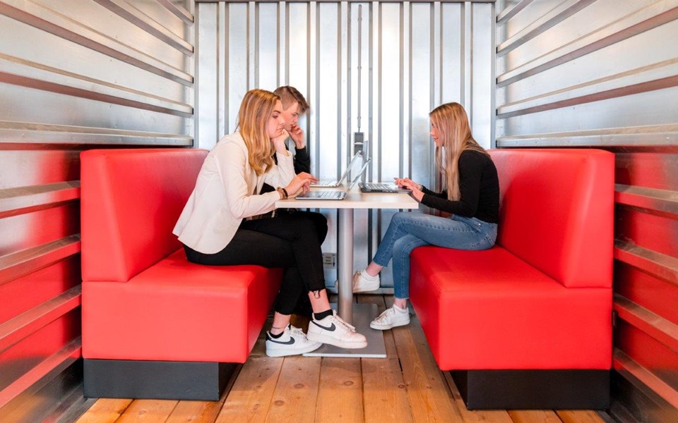 Drie studenten mediavormgeving zitten op twee rode bankjes met hun laptop op tafel 