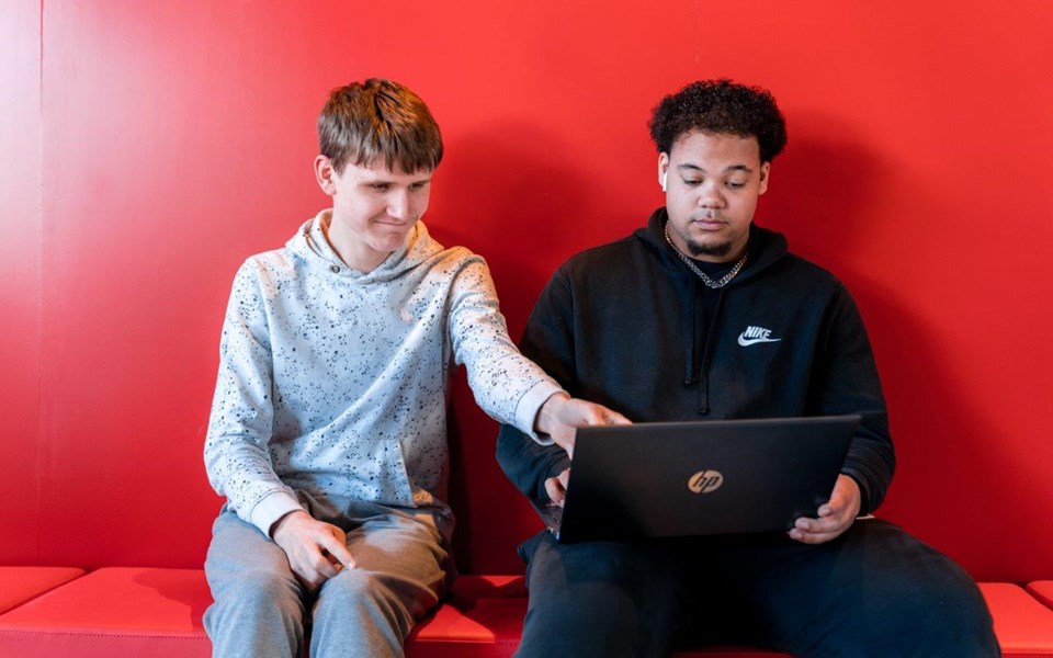 Twee studenten Medewerker ICT Support zitten op grote rode bank en kijken samen op één laptop