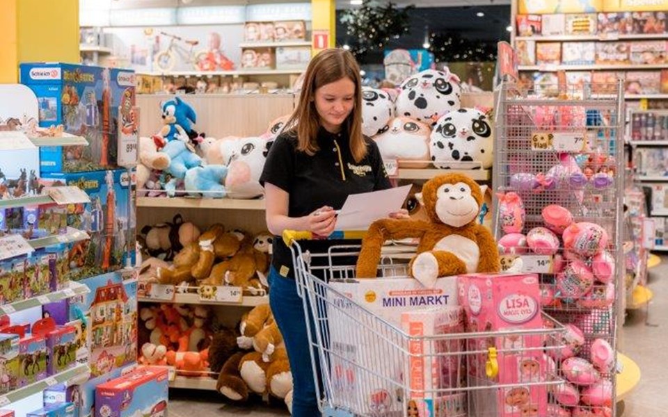 Vrouw rijdt met winkelwagen door speelgoedwinkel en kijkt op papier naar bestelling.
