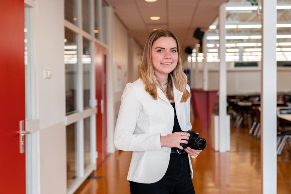 Mediavormgever, Iris Sijbom 2022 (16) Student mediavormgeving staat met fotocamera om de nek in grote ruimte met stoelen