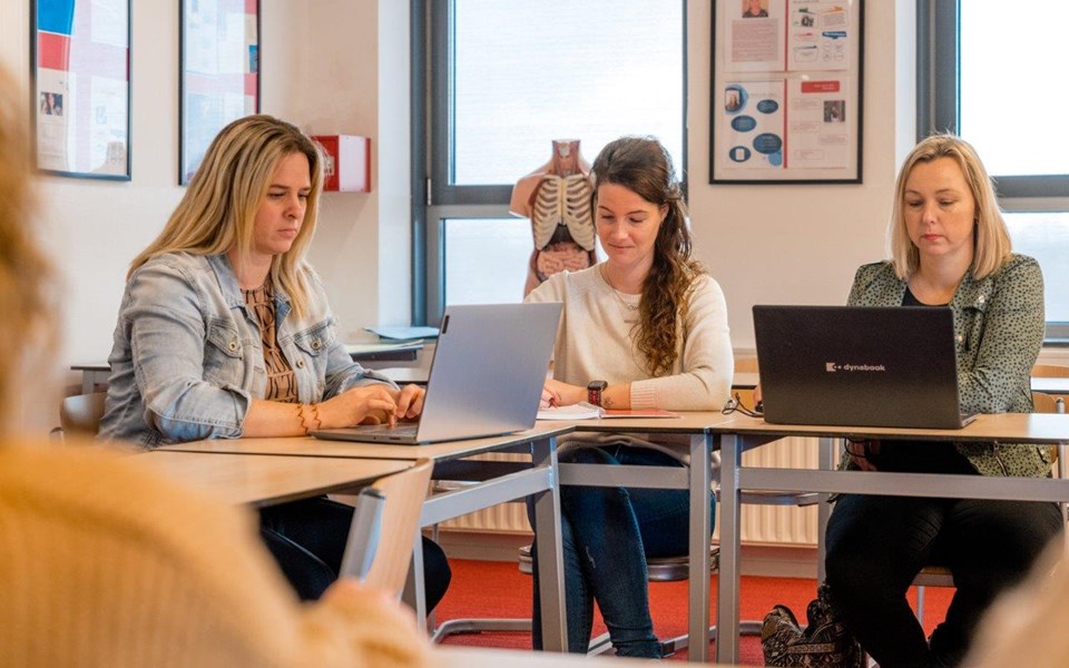 Drie volwassenen studenten verpleegkunde werken op laptop in klaslokaal.