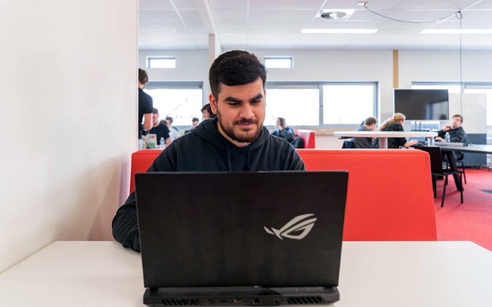 Student Software Developer achter de laptop met op de achtergrond lokaal vol andere studenten