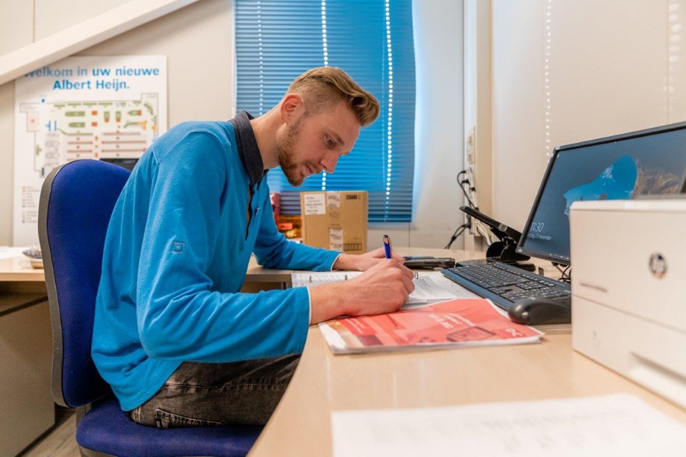 Man op kantoor zit achter computer en schrijft iets op papier.